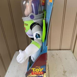 May include: A Buzz Lightyear plush toy from Disney's Toy Story, fully poseable. The toy is in its original packaging, featuring a clear helmet, white spacesuit with green and purple accents. The packaging includes the text "BUZZ LIGHTYEAR" and "FULLY POSEABLE!"