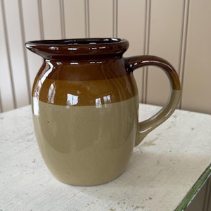 May include: A ceramic pitcher with a handle, featuring a two-tone design. The top section is a dark brown, transitioning to a lighter brown band, and then a beige base. The pitcher has a spout for easy pouring and is suitable for serving beverages.