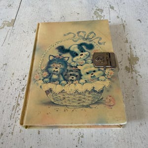 May include: A rectangular diary with a cream-colored cover featuring a printed illustration of four puppies sitting inside a woven basket with a blue ribbon. The diary includes a silver-toned metal locking mechanism with a heart-shaped keyhole.