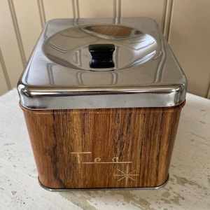 May include: A vintage tea tin with a stainless steel lid and a faux wood grain body. The lid has a black handle. The word "Tea" is printed on the front in a stylized font, with a starburst design below.