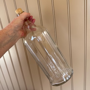 May include: A clear glass bottle with a cork stopper. The bottle has a rectangular shape with vertical grooves and is held by a hand. The bottle is empty and appears to be for decorative purposes.