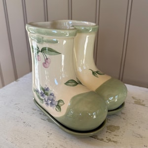 May include: Cream-colored ceramic boot-shaped planter with a light green rim and toe. The planter is decorated with hand-painted purple flowers, green leaves, and pink cherries. The planter sits on a matching green base.