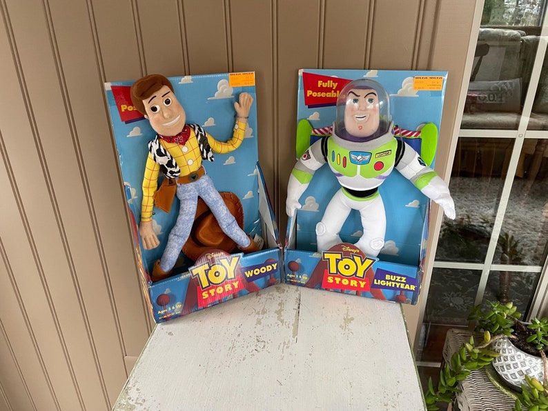 May include: Two Toy Story character plush toys, Woody and Buzz Lightyear, in their original boxes. Woody is in a cowboy outfit, and Buzz is in a spacesuit. The boxes have the Toy Story logo and are set against a light-colored background.