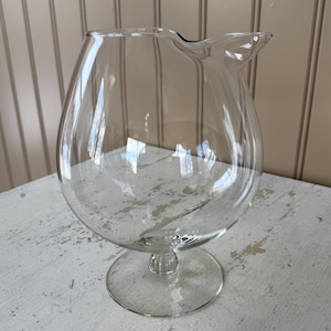 May include: A clear glass pitcher with a rounded body and a spout. The pitcher sits on a circular base and has a stem. The glass is transparent, allowing a view of the interior. The pitcher is likely used for serving beverages.