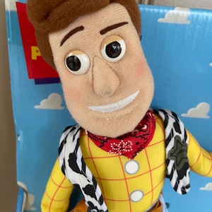 May include: A plush Woody doll from the Toy Story franchise. The doll features a yellow plaid shirt, blue jeans, a red bandana, and a cow-print vest. The doll has a stitched smile and large, expressive eyes. The packaging is visible in the background.