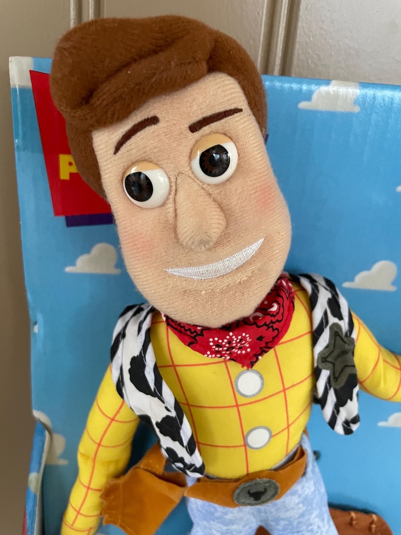May include: A plush Woody doll from the Toy Story franchise. The doll features a yellow plaid shirt, blue jeans, a red bandana, and a cow-print vest. The doll has a stitched smile and large, expressive eyes. The packaging is visible in the background.