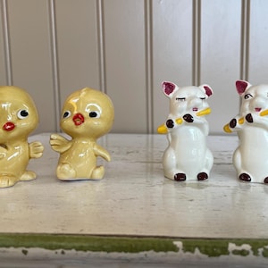 May include: A collection of ceramic figurines, including two yellow chicks with red beaks and two white pigs with yellow instruments. The figurines are arranged on a white and green surface.