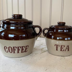 May include: Two ceramic canisters, one large and one small, with lids and handles. The larger canister is labeled "COFFEE" and the smaller one is labeled "TEA". Both are brown and beige.