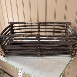 May include: A rustic, rectangular basket crafted from dark brown wooden sticks, tightly woven together. The basket has a sturdy, open design, perfect for storage or display. The basket is sitting on a white and green table.
