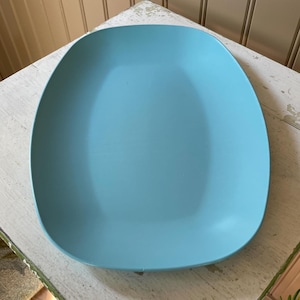 May include: A light blue, oval-shaped serving platter. The platter has rounded corners and a smooth surface. The color is a solid, muted blue. The platter is sitting on a white, distressed wooden surface.