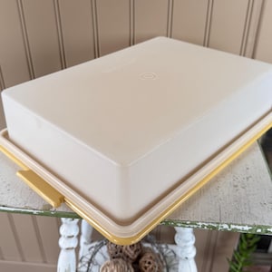May include: A rectangular, beige plastic container with a yellow rim and handle. The lid is flat and has a slightly rounded edge. The container is sitting on a distressed white table.