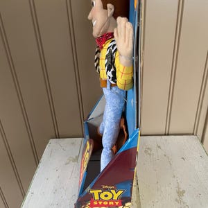 May include: A plush Woody doll from Disney's Toy Story, in its original packaging. Woody is wearing a yellow shirt with red stitching, a cow-print vest, blue jeans, and a red bandana. The packaging features the Toy Story logo and the words "Woody" and "Fully Poseable!"