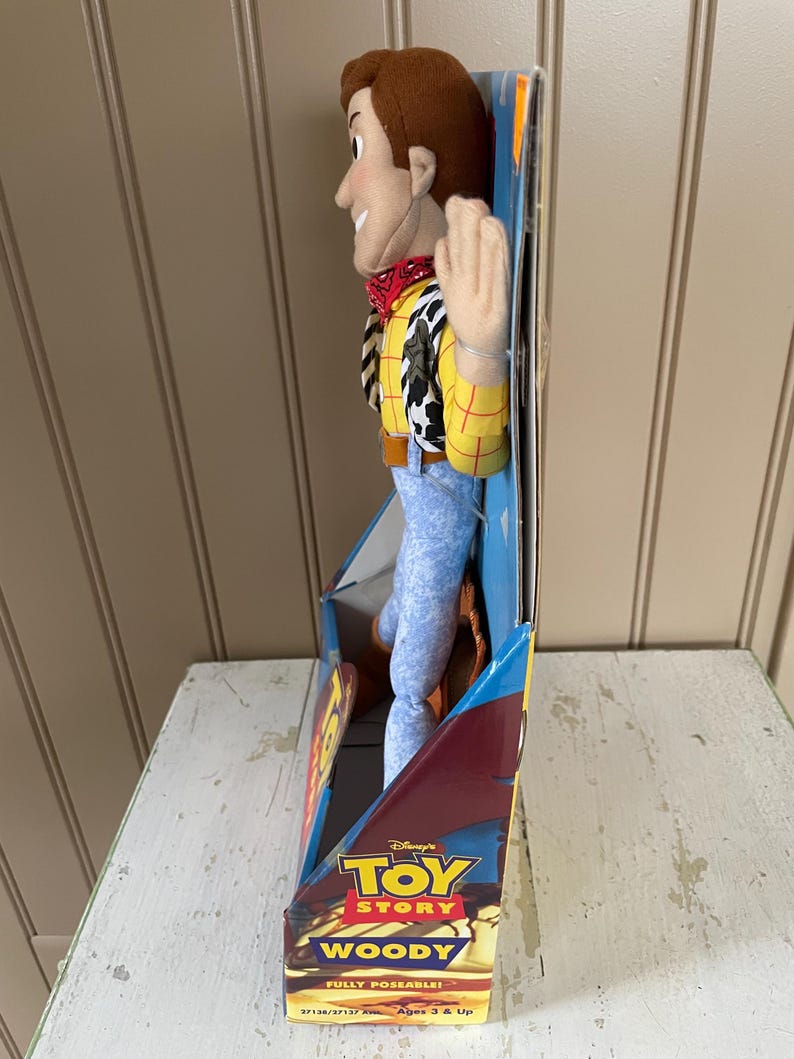 May include: A plush Woody doll from Disney's Toy Story, in its original packaging. Woody is wearing a yellow shirt with red stitching, a cow-print vest, blue jeans, and a red bandana. The packaging features the Toy Story logo and the words "Woody" and "Fully Poseable!"