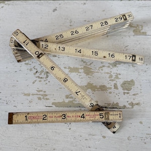 May include: An antique folding ruler, featuring a cream-colored finish with black markings. The ruler is marked with numbers and measurements in inches, with the "STANLEY" brand visible. The ruler is partially open, displaying its segmented design.