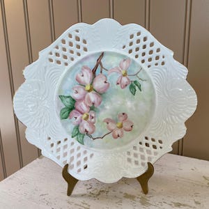 May include: A white, decorative plate with a floral design. The plate features a central painting of pink dogwood blossoms on a branch, surrounded by a white, openwork border. The plate is displayed on a gold-colored stand.