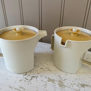 May include: Two white plastic pitchers with yellow lids and a handle. The pitchers are designed for pouring liquids like milk or cream.