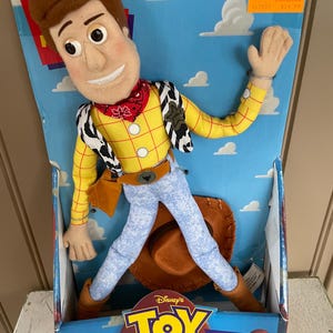 May include: A Woody doll from Disney's Toy Story, in its original box. The doll is wearing a yellow shirt, blue jeans, a red bandana, and a cowboy hat. The box has the Toy Story logo and the words "Woody".