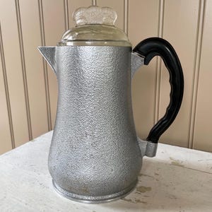 May include: Vintage silver-toned coffee pot with a textured surface and a black handle. The glass lid has a decorative knob. The coffee pot is designed with a spout for easy pouring. A classic kitchenware item.
