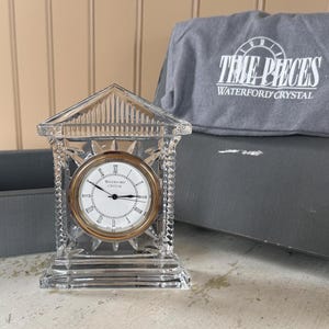 May include: A clear crystal clock with a gold-tone bezel and Roman numeral hour markers. The clock is in a triangular shape and has the words "WATERFORD CRYSTAL" on the clock face. A gray bag with the words "TIME PIECES WATERFORD CRYSTAL" is behind the clock.