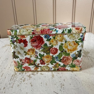 May include: A rectangular metal storage tin featuring a vibrant all-over floral pattern with red, yellow, pink, and white roses and green leaves against a white background. The container has a matching lid and a thin gold-toned metal rim.