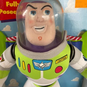 May include: Buzz Lightyear plush doll with a clear plastic helmet. The doll has a green and white spacesuit with the word "LIGHTYEAR" on the chest. The doll is in a box with the words "Fully Posed" visible.