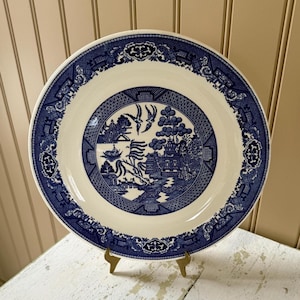 May include: A round, white and blue Willow pattern plate. The plate features a detailed blue design around the rim and a central scene with a pagoda, birds, and a bridge. The plate is displayed on a gold-colored stand.