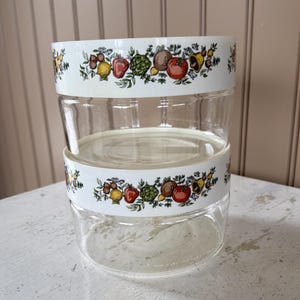 May include: Two clear glass food storage containers stacked on top of each other. Each container has a white band around the top with a colorful fruit and vegetable design. The containers are sitting on a white surface.