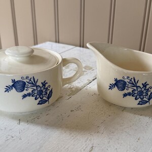 May include: A cream-colored ceramic sugar bowl with a matching lid and a cream-colored ceramic creamer pitcher. Both pieces feature a blue floral pattern with a central flower and a bud on a stem, each equipped with a single side handle.