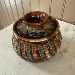 May include: A vintage Dryden Potter's Wheel Original ceramic bowl with a lid. The bowl is brown with green and tan accents. The lid has a raised center and floral designs. The bowl has a textured, ribbed design around the base.