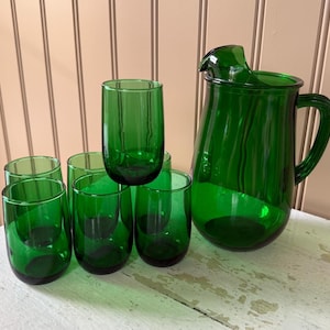 May include: A vintage green glass pitcher and six matching tumblers. The pitcher has a handle and a spout. The glasses are cylindrical. The set is made of a deep emerald green glass, perfect for serving beverages.