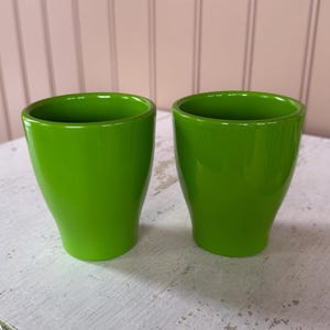 May include: Two bright green ceramic planters. The planters have a glossy finish and a slightly flared shape. They are empty and ready for planting. The background is a light pink and white.