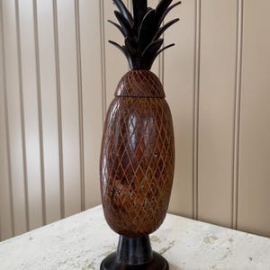 May include: A decorative pineapple-shaped container with a dark brown base and top, and a textured, diamond-patterned body. The pineapple top is dark brown, and the body is a lighter brown. The container is on a white surface.
