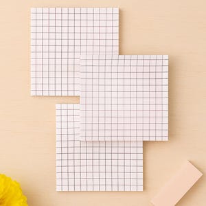 Pode incluir: Três blocos de notas quadrados brancos com um padrão de grade estão dispostos sobre uma superfície de madeira clara. Uma pequena nota autocolante retangular rosa claro e uma flor amarela também estão presentes. Ideal para tomar notas.