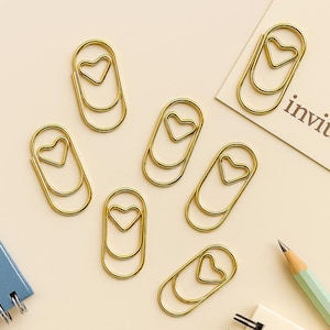 May include: Gold-colored paper clips with a heart-shaped design. The clips are scattered on a light beige surface, alongside a notepad, pencil, and a card with the word "invit" printed on it. The clips are a decorative and functional office supply.