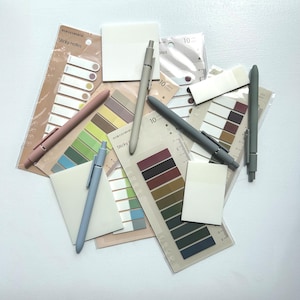 Puede incluir: Un surtido de artículos de papelería, que incluyen bolígrafos, notas adhesivas y pestañas adhesivas. Los bolígrafos son de varios colores, como rosa, gris y azul. Las notas adhesivas y las pestañas vienen en una variedad de colores y tamaños.