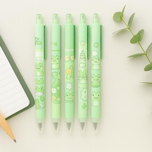 May include: Five green pens with frog illustrations. Each pen features a different frog design, with additional details like flowers and clouds. The pens are arranged vertically, with the tips pointing down. The word "Fickle" is printed on one of the pens.