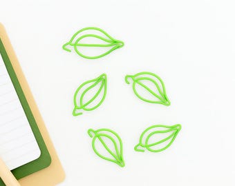 May include: Five bright green leaf-shaped paper clips are scattered on a white surface. A notepad with lined paper and a tan and green cover is in the upper left corner. The clips are made of wire.