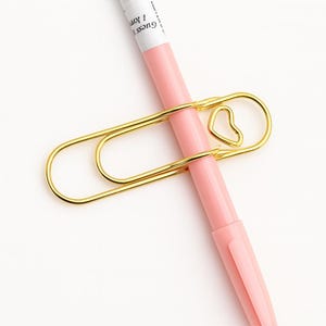 Puede incluir: Un clip de papel dorado con un diseño en forma de corazón que sujeta un bolígrafo rosa. El bolígrafo tiene texto blanco que dice "Guess 1 Jon". El clip de papel es de metal y tiene un acabado brillante. El bolígrafo es de color rosa claro.