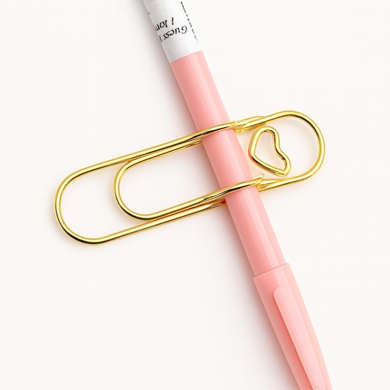 May include: A gold-colored paperclip with a heart-shaped design holding a pink pen. The pen has white text that reads "Guess 1 Jon". The paperclip is made of metal and has a shiny finish. The pen is a light pink color.