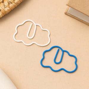 May include: Two cloud-shaped paper clips, one white and one blue, sit on a tan surface. The clips have a wavy outline and a straight, curved section. A woven basket and a small notebook are in the background.