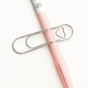 May include: A silver metal paperclip holding a pink pen. The paperclip has a heart-shaped design element. The pen has white text on it.