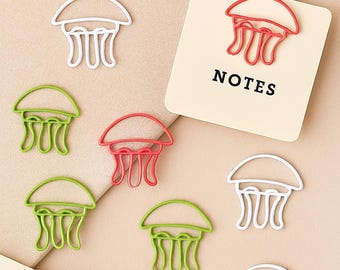 May include: A collection of colorful jellyfish-shaped paper clips in red, green, and white. A small notepad with the word "NOTES" is visible. The paper clips are arranged on a light brown surface.
