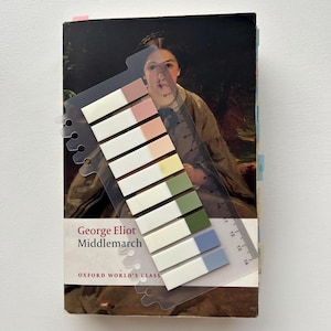 Op de afbeelding: Een exemplaar van "Middlemarch" van George Eliot met een transparante liniaal en gekleurde plaknotities. Het boek heeft een portret op de omslag en het Oxford World's Classics logo. De plaknotities zijn in verschillende pastelkleuren.