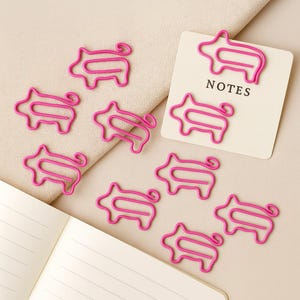 May include: A selection of vibrant pink pig-shaped paper clips. One clip is attached to a small white card with the word "NOTES" printed in black. The clips are scattered on a beige surface, near an open notebook.