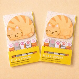 以下が含まれることがあります： 2つの猫をテーマにした付箋のパック。各パックには、眠っているオレンジ色のトラ猫の形をしたメモパッドと足の形をした付箋が付いています。「stick & bigmemo」というテキストがパッケージに印刷されています。