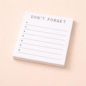 Peut inclure: Un bloc-notes blanc avec les mots "DON'T FORGET" imprimés en noir en haut. Le bloc-notes comporte des lignes horizontales et des points pour une liste de contrôle. Le bloc-notes est sur une surface rose clair.