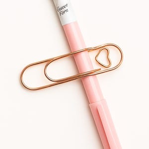May include: A gold-colored paperclip with a heart-shaped design, resting on a pink pen. The pen has the text "Gwet Forn" printed on it. The paperclip is a decorative office supply.