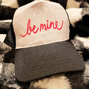 May include: A white and black trucker hat with a red cursive inscription that says "be mine".