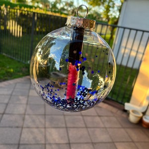 Może przedstawiać: Przezroczysta szklana ozdoba choinkowa wypełniona niebieskim brokatem i różową szminką w sztyfcie.