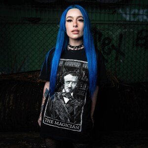 May include: A woman with bright blue hair is wearing a black t-shirt with a white graphic of a man with a raven on his shoulder. The graphic is labelled "The Magician".